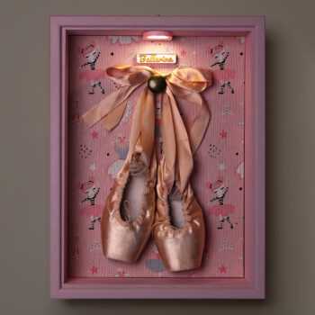Caja para puntas de ballet Clara iluminada en un ambiente oscuro, con marco rosa y fondo estampado de bailarinas